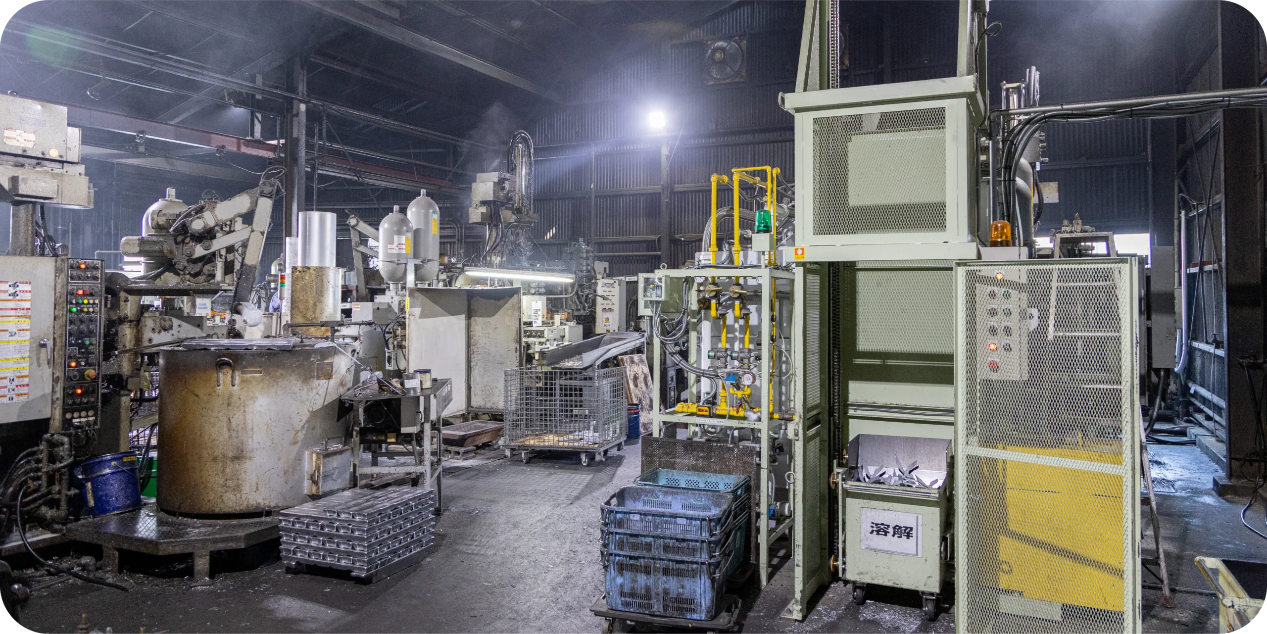 Wide view of a metal casting factory interior with melting furnaces, machinery, and parts bins.