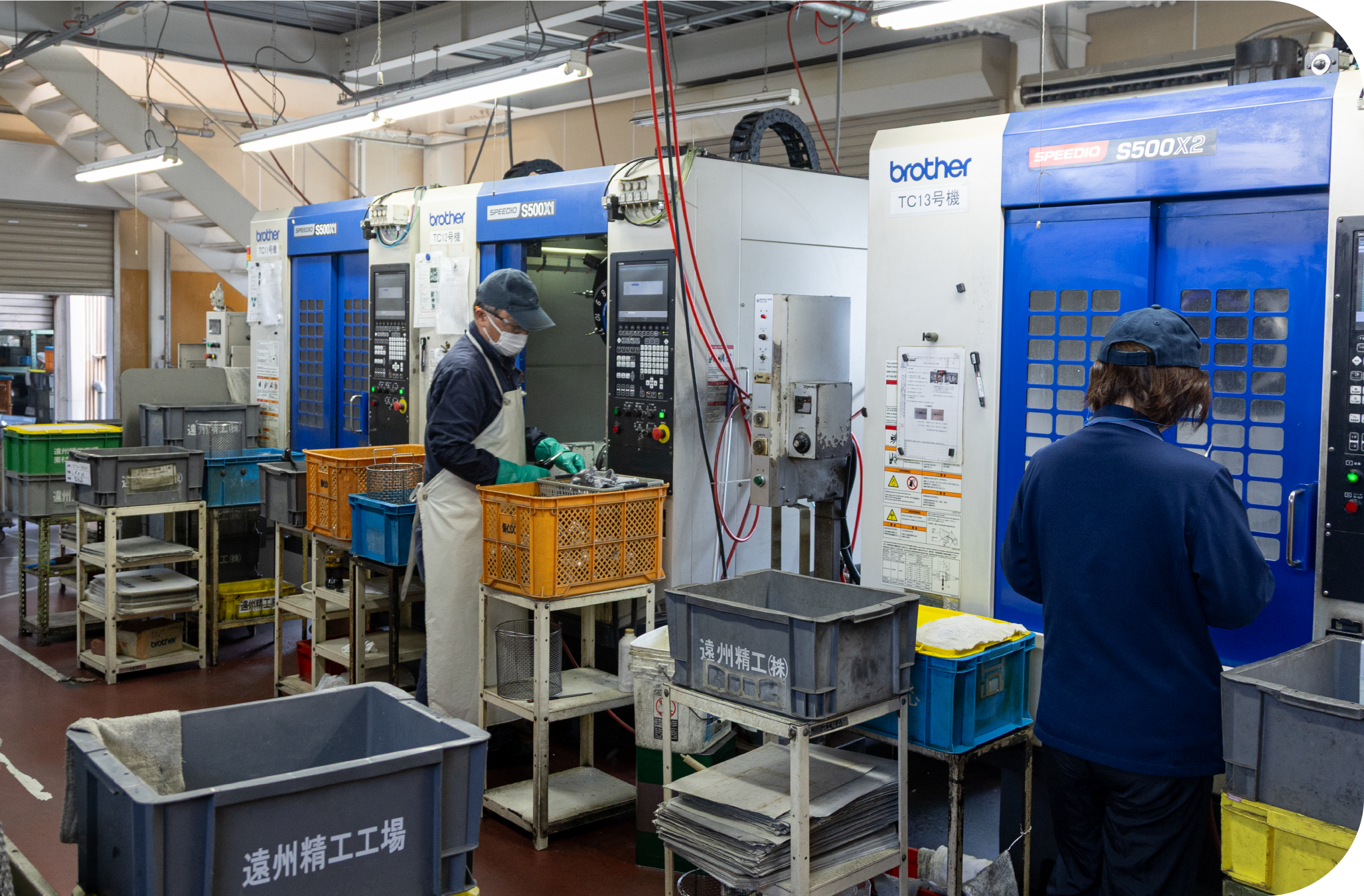 Workers operating a row of CNC machining centers with parts bins on a factory floor.