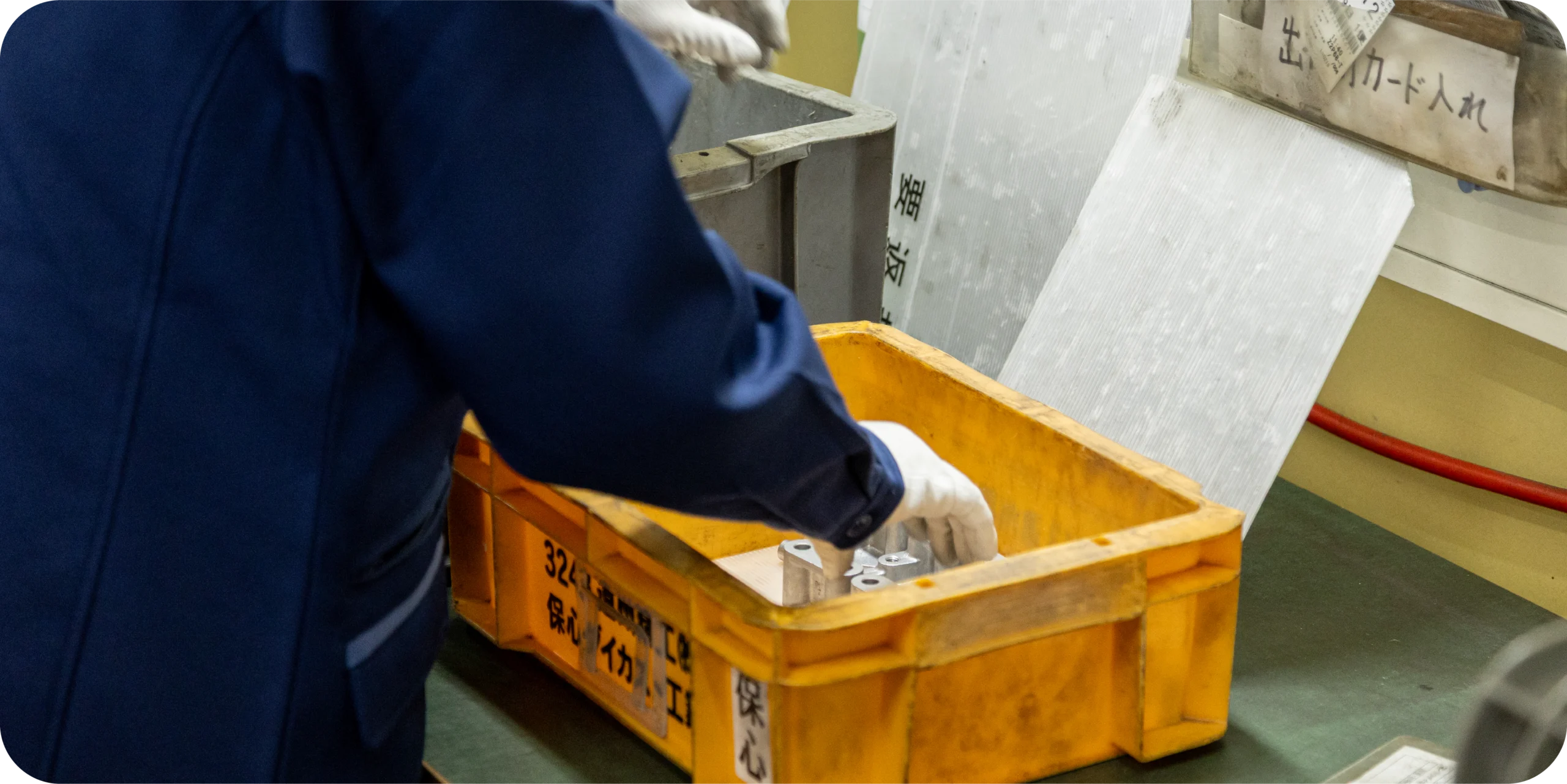 Gloved worker placing machined metal parts into a labeled bin at a workstation.