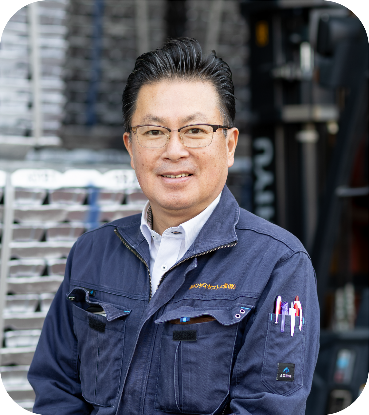 Smiling man in a work jacket standing in front of stacked metal ingots at a factory.